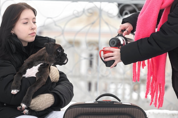 two female travelers with a dog drink a hot drink from a thermos on a winter day