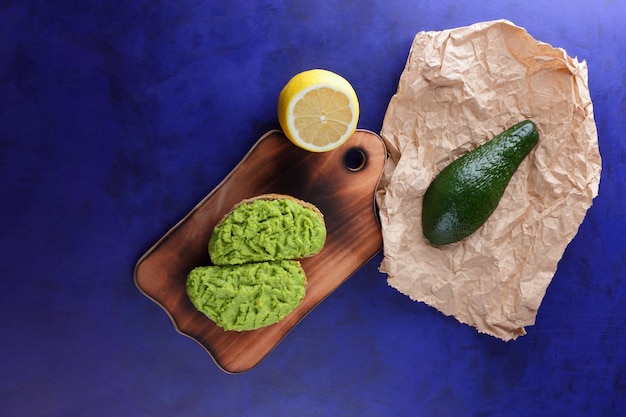 Two fresh green avocado toasts on a wooden board Toasts half an avocado and a yellow lemon in a peel on a blue background Closeup