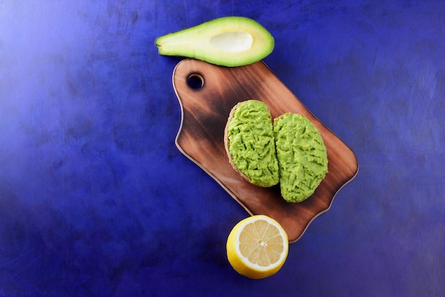 Two fresh green avocado toasts on a wooden board Toasts half an avocado and a yellow lemon in a peel on a blue background Closeup