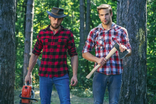 Two friends lumberjack worker in the forest woodcutter with axe and lumberjack with chainsaw handsom