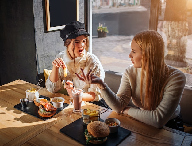 Two friends sit together inside at table. Young woman in cap argue with another. She is mad. Blonde girl look at her friend and talk to her.