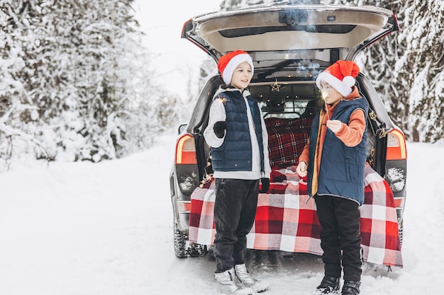 Two friends teenagers boys in red santa hats with with sparkles Bengal fire standing near trunk of car decorated for Christmas and New Year in snowy winter forest Road trip and local travel