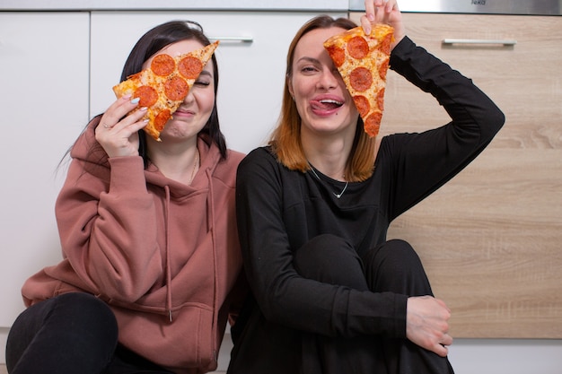 Two gay girls joking apply pieces of pizza to their face while sitting on the kitchen floor near the cupboards high quality photo