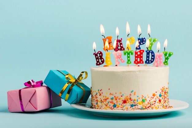 Two gift boxes near the cake with happy birthday candles against blue backdrop