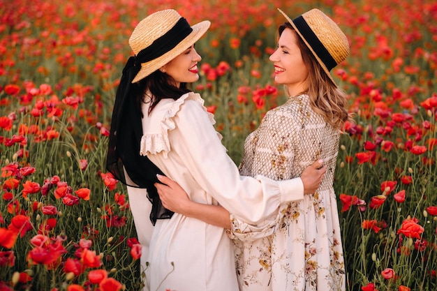 Two girlfriends in dresses and a hat in a poppy field in summer at sunset