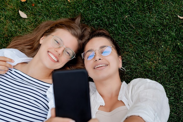 Two girlfriends student take a photo selfie on mobile phone lying on the green grass