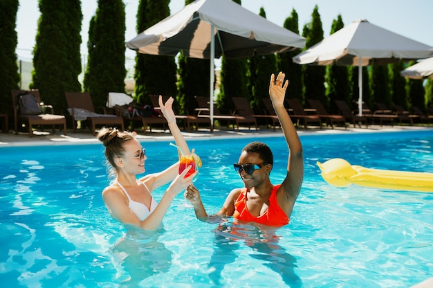 Two girlfriends with coctails swim in the pool