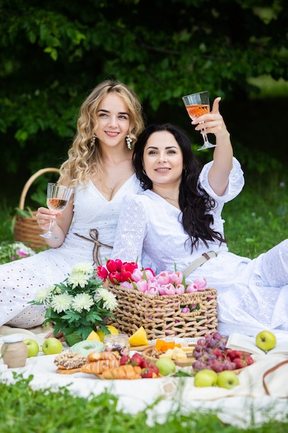 Two girls are resting in park sitting on a picnic blanket with fruits and wine