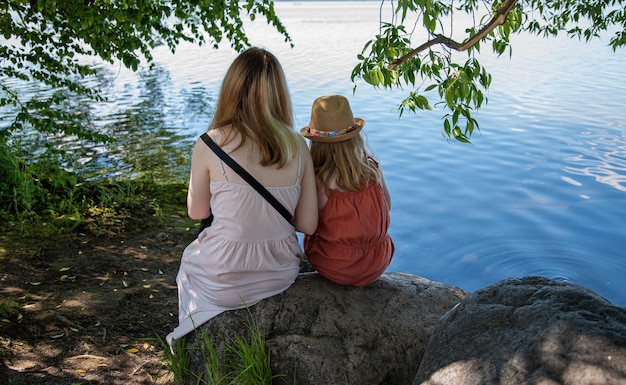 Two girls are sitting on a rock under a tree on the shore of a lake on a sunny summer day Without face