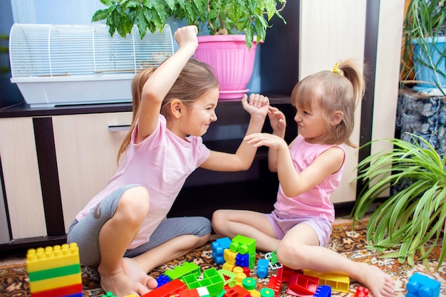 Two girls sisters are sitting at home on the floor and playing in a plastic construction set.