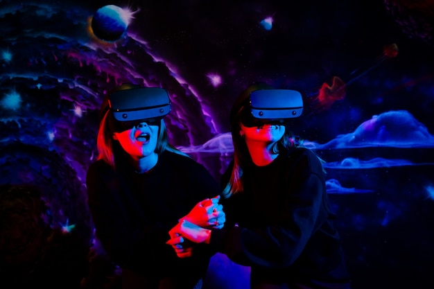 Two girls sisters friend with virtual glasses holding hands are having fun in the game room in neon blue and red light