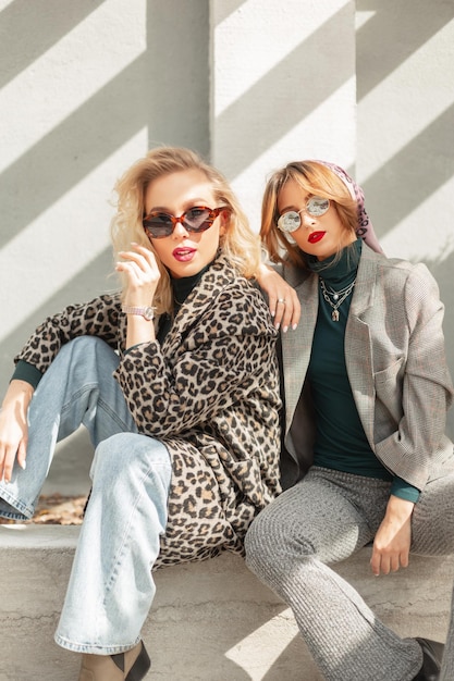 Two glamorous young girls in fashionable clothes with sunglasses are sitting on the street