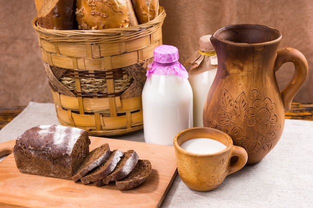 Two glass bottles of milk between a basket of bread and a wooden jug and cup beside a cutting board of sliced pumpernickel