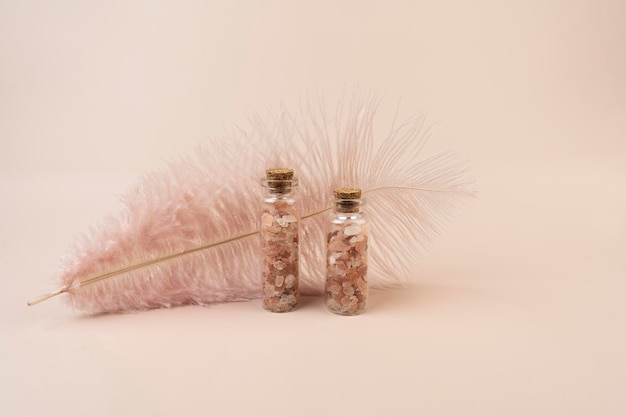 Two glass bottles with pink salt for spa procedures standing on a pink background a pink feather behind them