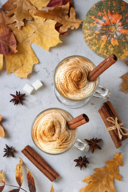 Two glass cups pumpkin latte with spices on grey background with pumpkins and autumn leaves