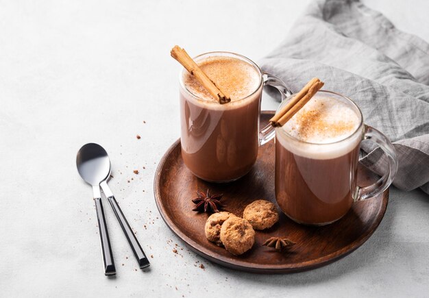 Two glass mugs with hot chocolate and milk foam on a wooden board on a gray background with cinnamon sticks anise star cookies and spoon