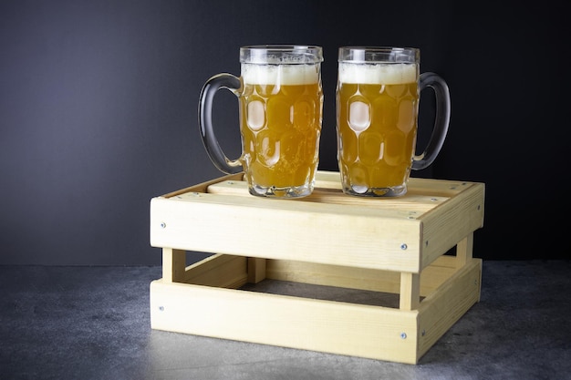 Photo two glass mugs with light beer on wooden box on gray table against black background