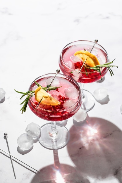 Two glass of pomegranate iced cocktail with rosemary and lemon on white marble background isolated with hard shadows text space Festive drink valentines day concept