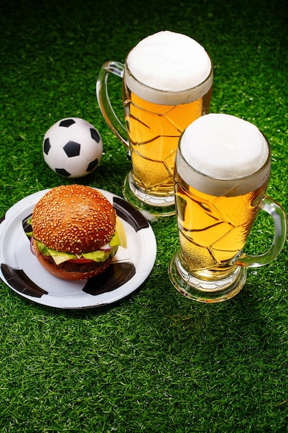 Two glasses of beer hamburger and soccer ball on green grass