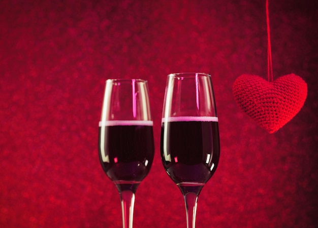 Two glasses of champagne or sparkling wine on a red bokeh background, a red knitted heart. Valentine's Day Concept