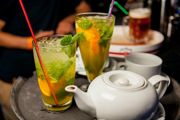 Two glasses of cocktails with mint, and a teapot with tea, stand on the bar.