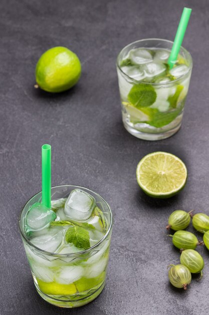 Two glasses of cold drink with lime, mint and ice. Straw in glass. Slices of lime and gooseberries on table. Black background. Top view