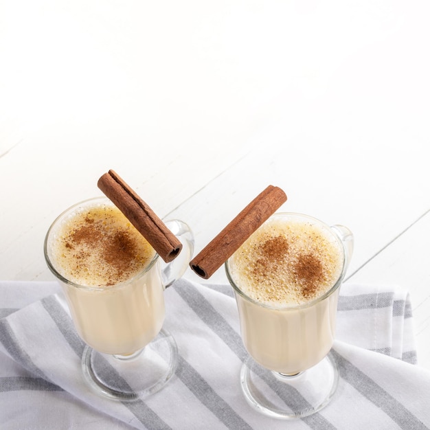 Two glasses of traditional eggnog with cinnamon sticks on striped napkin on white wooden table