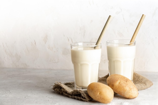 Two glasses of vegan potato milk with bamboo drinking straws and potato tubers on neutral backdrop