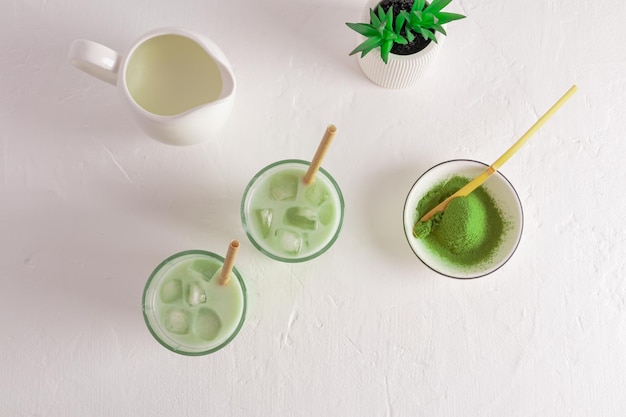 Two glasses with green iced tea matcha latte a bowl of matcha powder a milk jug with milk top view cooking white background wellness drink