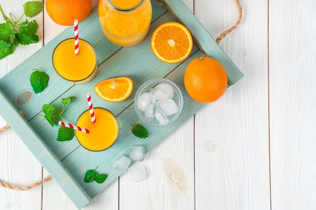 Photo two glasses with juice and tubes, oranges, mint and ice cubes on a board on a light desk. top view with copy space.