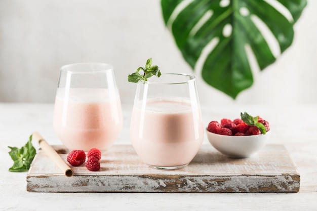 Two glasses with a Lassi drink with raspberries, mint and a bamboo tube on a white wooden board. Light background with a monstera leaf. Horizontal orientation. Indian traditional refreshing drink.