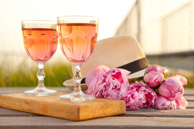 Two glasses with rose wine and pink peony bouquet refreshing drink blossoming flowers and mens hat