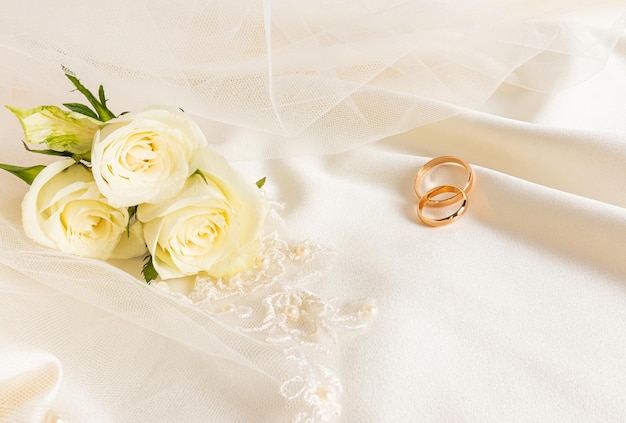 Two gold wedding rings leaned on the folds of satin beige fabric with a bouquet of white live roses wedding background cover postcard