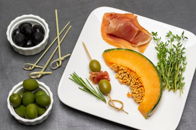 Two green olives and jamon on skewer Slice of melon and sprigs of thyme on plate Black and green olives in bowls