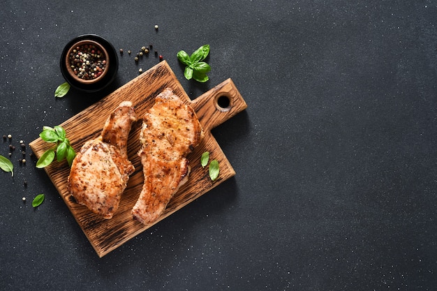 Two grilled pork steaks with garlic and basil on a wooden board on a black background with space for the text. View from above.