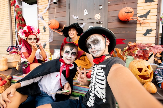 Two halloween boys making selfie while sitting on staircase on background of cute girl and happy young woman in witch hat and yellow dress