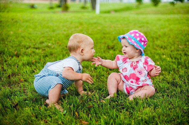 Two happy baby boy and a girl age 9 months old, sitting on the grass and interact, talk, look at each other.