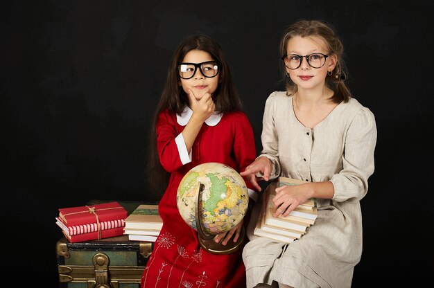 two happy beautiful schoolgirls with globe and books sitting on chest on a black backgroud