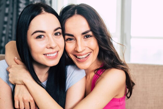 Two happy beautiful young smiling girlfriends or sisters in bright casual clothes are sitting on the sofa at home