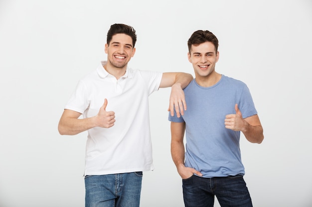 Two happy buddies 30s wearing casual t-shirt and jeans smiling and showing thumbs up on camera in studio, isolated over white background