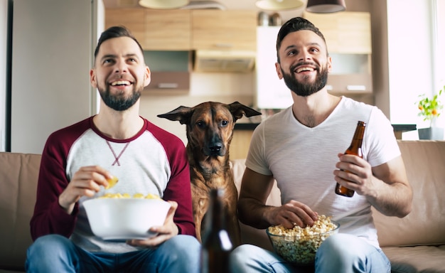 Two happy excited bearded friends watching TV or some sport match with dog while sitting on the couch at home on weekend