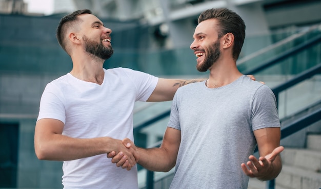 Two happy excited handsome bearded best friends in casual clothes are handshaking, hugging while standing outdoor.