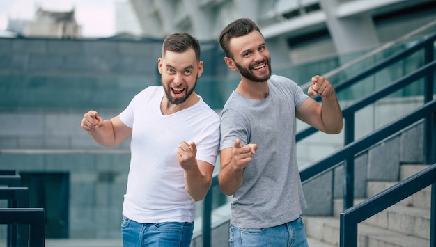 Two happy excited modern bearded best friends in casual clothes having fun and pointing on camera together outdoors.