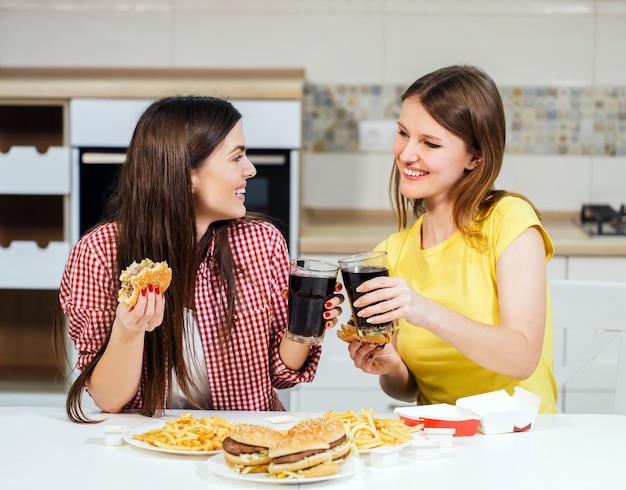 Two happy friends spending time together eating tasty fast food in home kitchen having nice pleasant talk