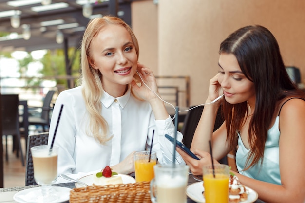 Two happy girls listening to music with shared earphones together in nice cafe