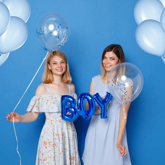 Two happy girls, young, in a studio, holding blue balloons and balloon with the inscription boy, blue background.