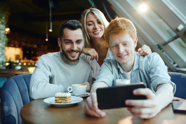 Two happy guys and pretty blonde girl making selfie on smartphone while gathered by table in cafe for rest and chat