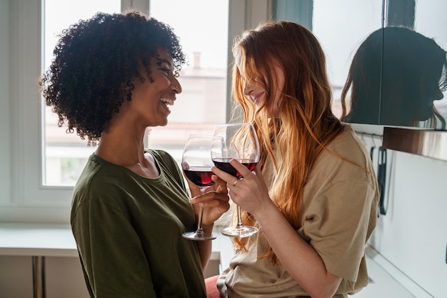 Two happy homosexual girls, in intimacy with each other, enjoy a glass of wine in the kitchen, looking at each other and smiling together.