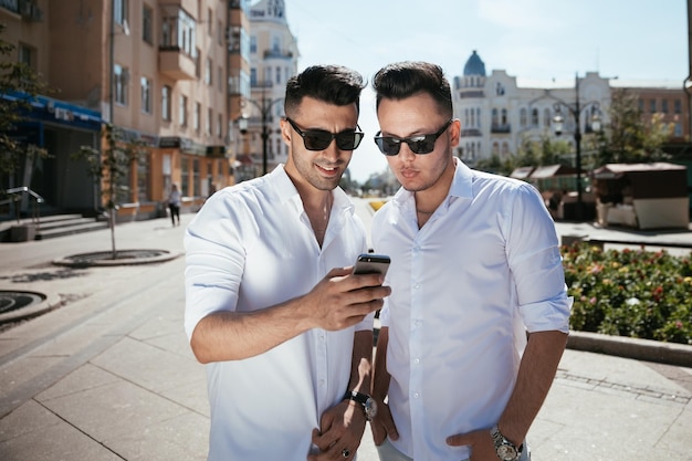 Two happy men gays laughing and watching into the phone on the street in the sunny day and discussing comething interestiong