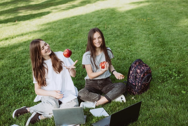 Two happy smiling female students are sitting in the park on the grass with books and laptops, eating apples, studying and preparing for exams. Distance education. Soft selective focus.